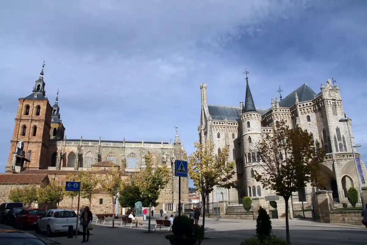A wide-angle photo shows a European city square with two large, ornate stone buildings, one with a reddish-brown tower and…