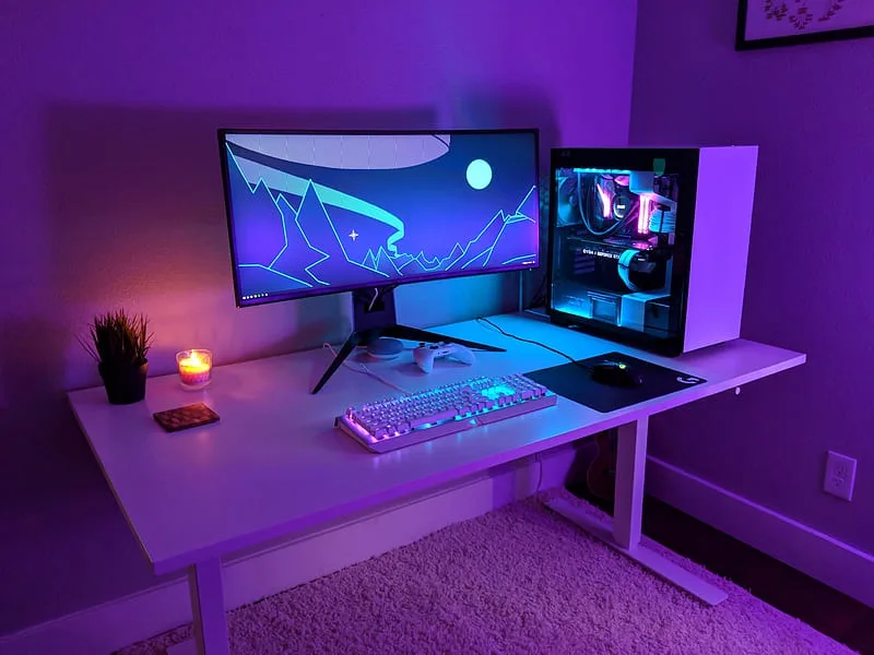 A well-lit, modern gaming setup with a large monitor displaying a stylized mountain scene, a glowing RGB keyboard, a gaming…