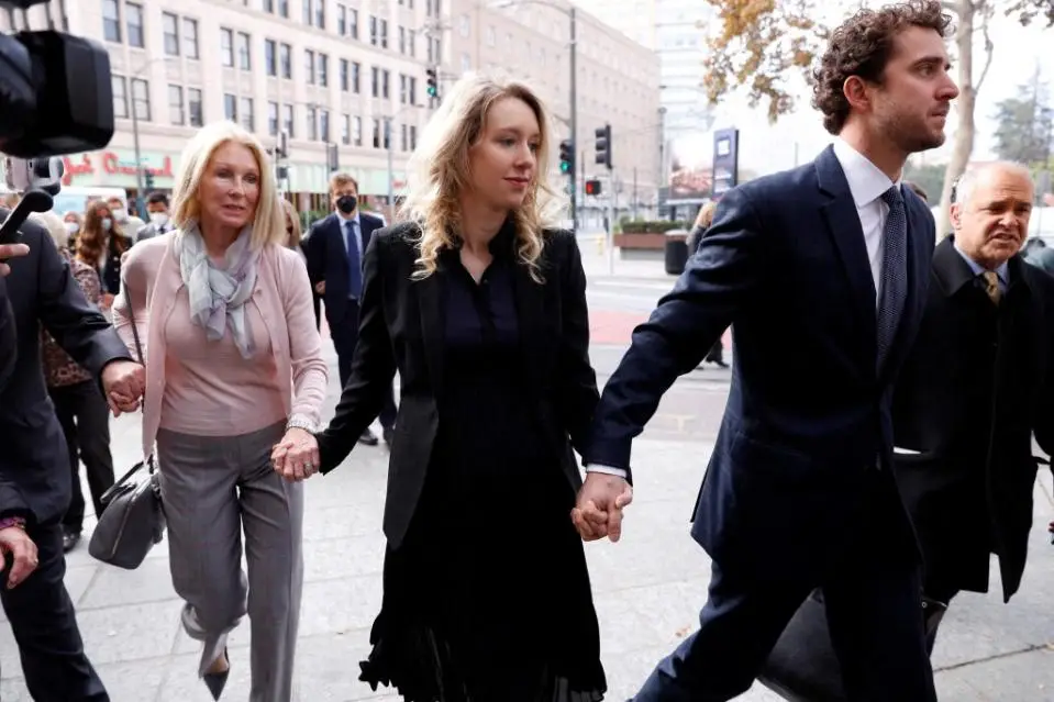 Three people walk down a city sidewalk holding hands, flanked by others and cameras. The central woman in a black dress…