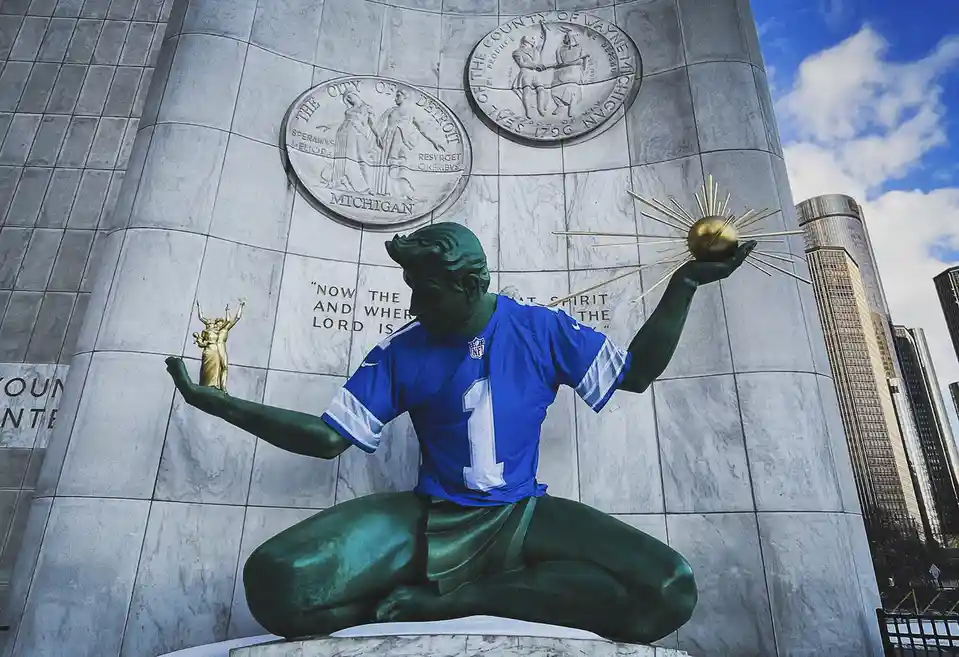 A green statue of a kneeling football player wearing a blue jersey with the number 1 and holding a golden trophy in one hand…