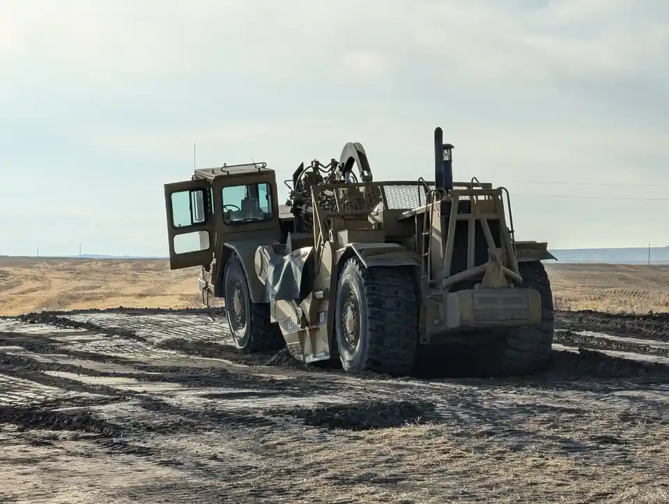 A large, tan-colored construction vehicle, possibly a Caterpillar loader, is stuck in deep mud in a barren field. Its…