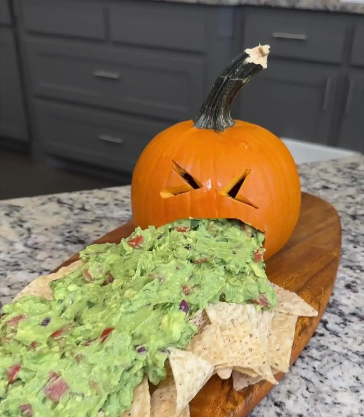 A carved orange pumpkin with a menacing face sits on a wooden board, its mouth filled with guacamole that spills over onto…