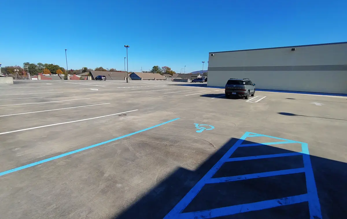 A wide-angle photo of an empty parking lot under a clear blue sky, with a single gray SUV parked near a building. In the…