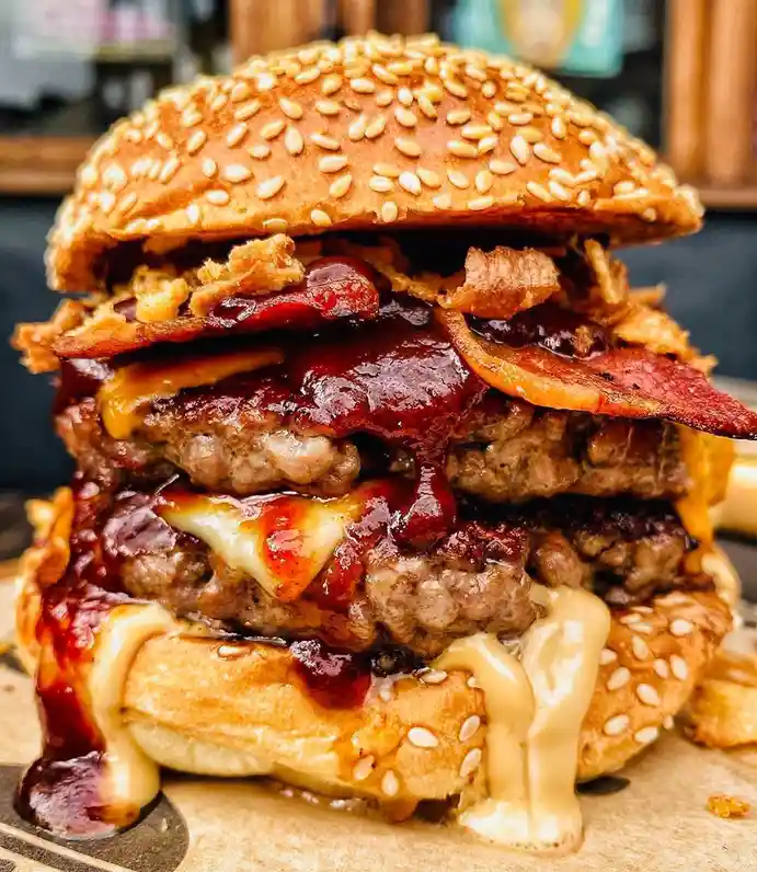 a towering, messy burger with multiple beef patties, melted cheese, crispy bacon, and a thick red sauce oozing out from…