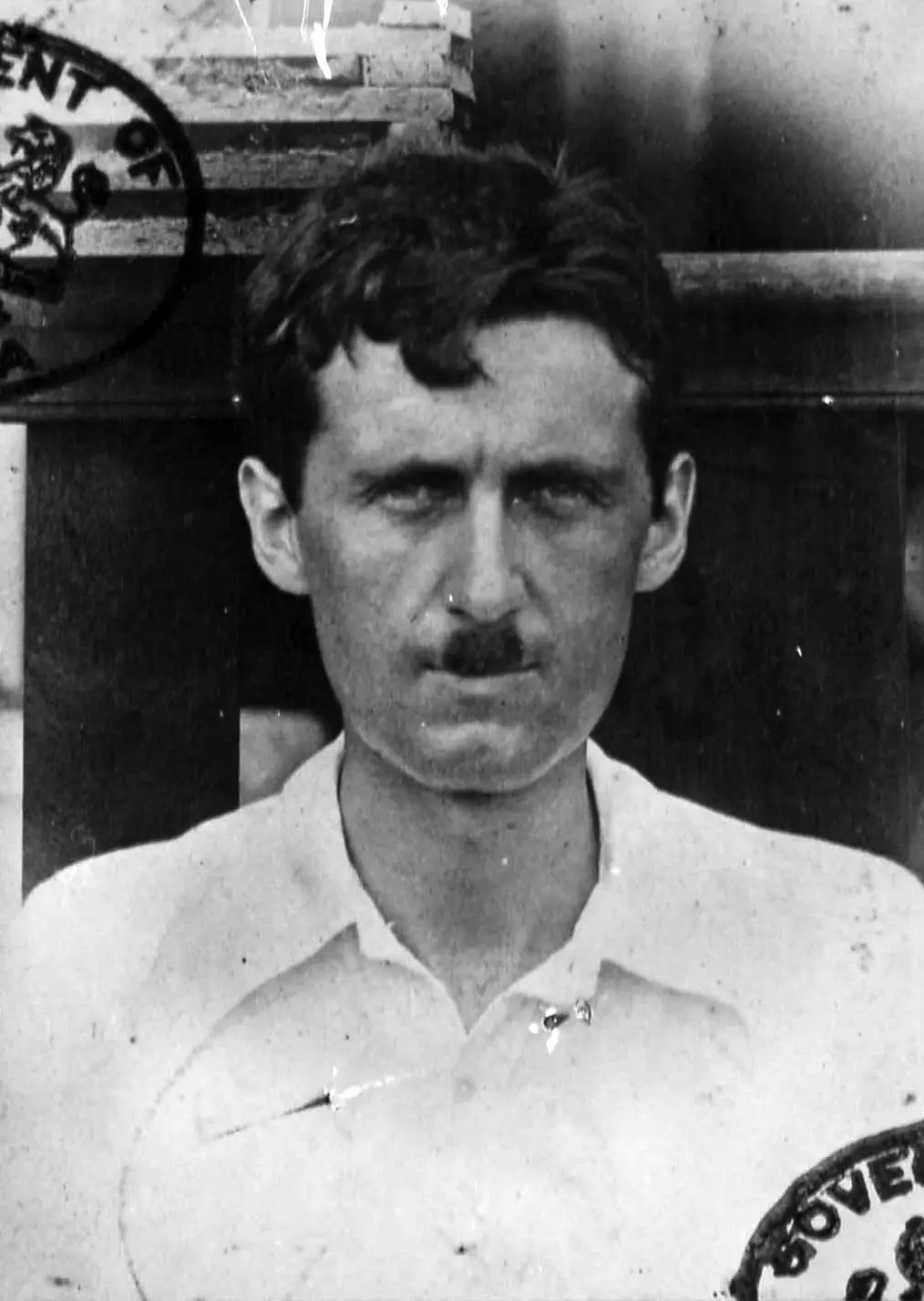 A black-and-white historical photograph shows a man with a mustache wearing a light-colored collared shirt with partial text…