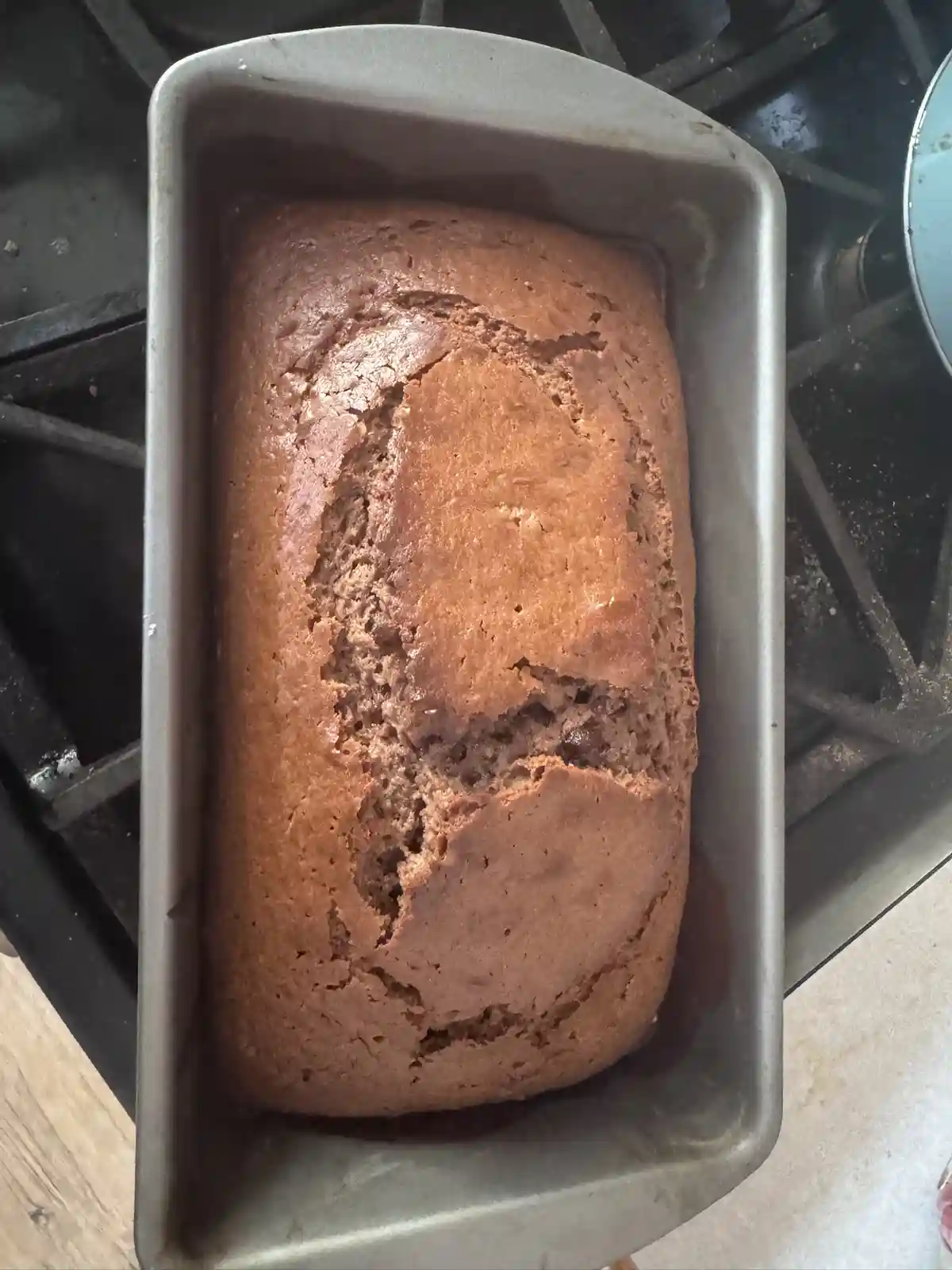 A freshly baked loaf cake sits in a metal loaf pan on a stovetop, its surface cracked and unevenly browned to resemble a…