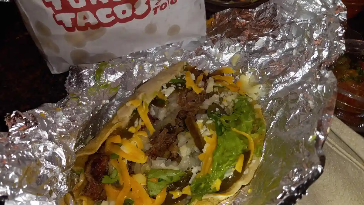 A close-up photo shows a large, messy taco wrapped in crinkled aluminum foil, overflowing with shredded cheese, guacamole…