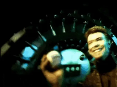 A split-screen image shows David Bowie in a dark, sci-fi setting holding a small, white, spherical robot. In the top row, he…
