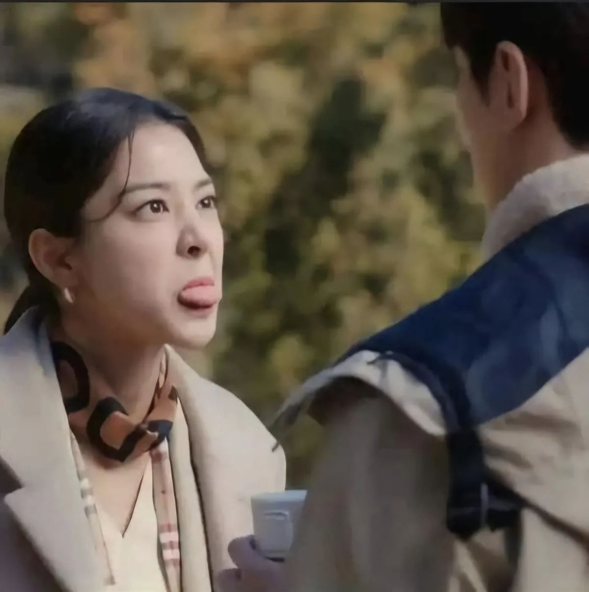 A woman with dark hair tied back, wearing a light-colored coat and a patterned scarf, looks up at a man with a surprised and…