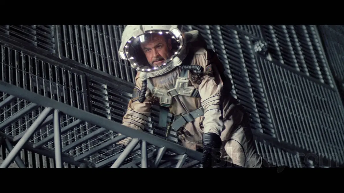 a man in a beige spacesuit with a reflective helmet is clinging to metal scaffolding, looking determined. The overlaid text…