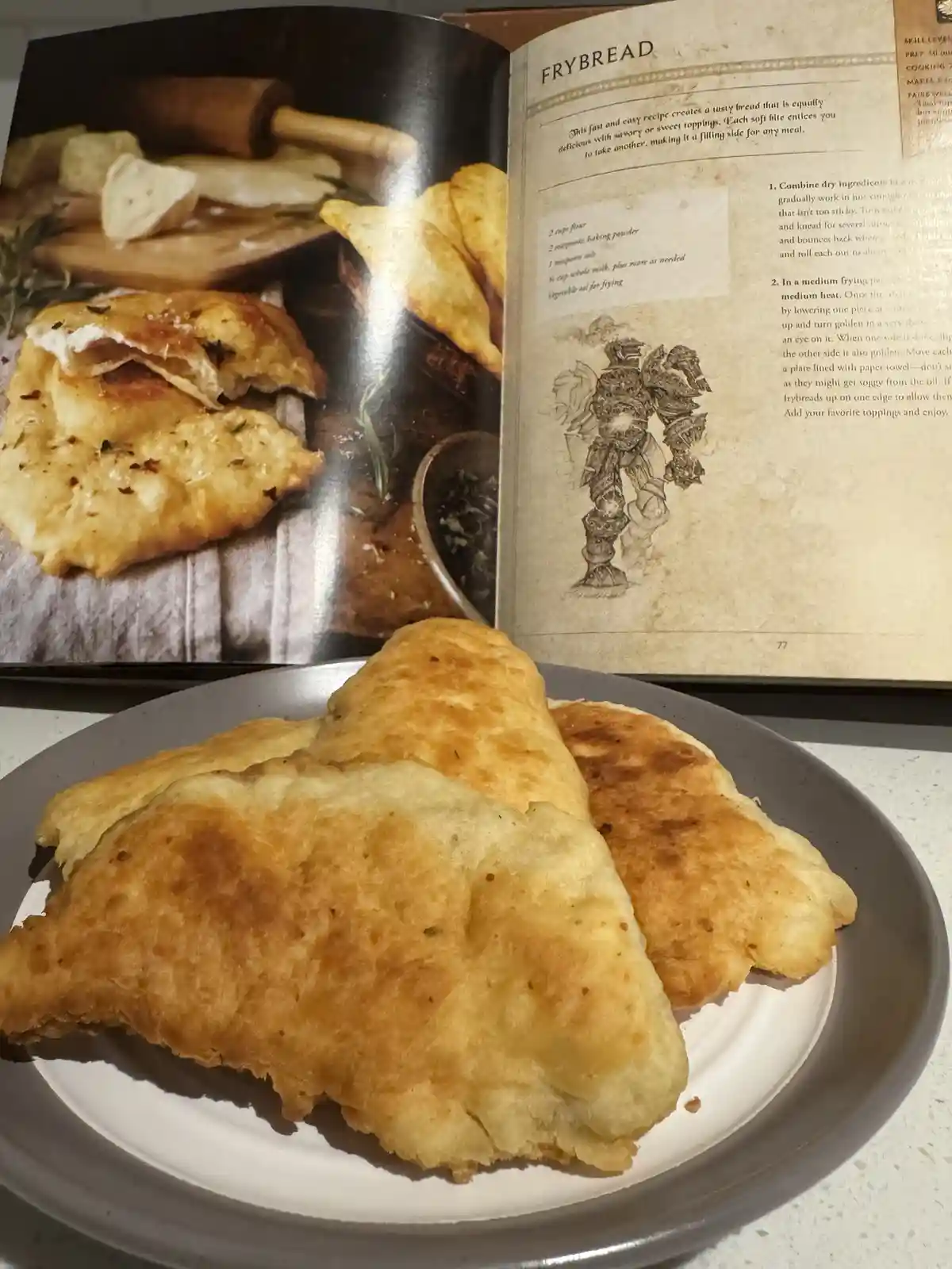 A plate of golden-brown, homemade frybread sits in front of an open cookbook. The cookbook page titled 'Frybread' shows a…