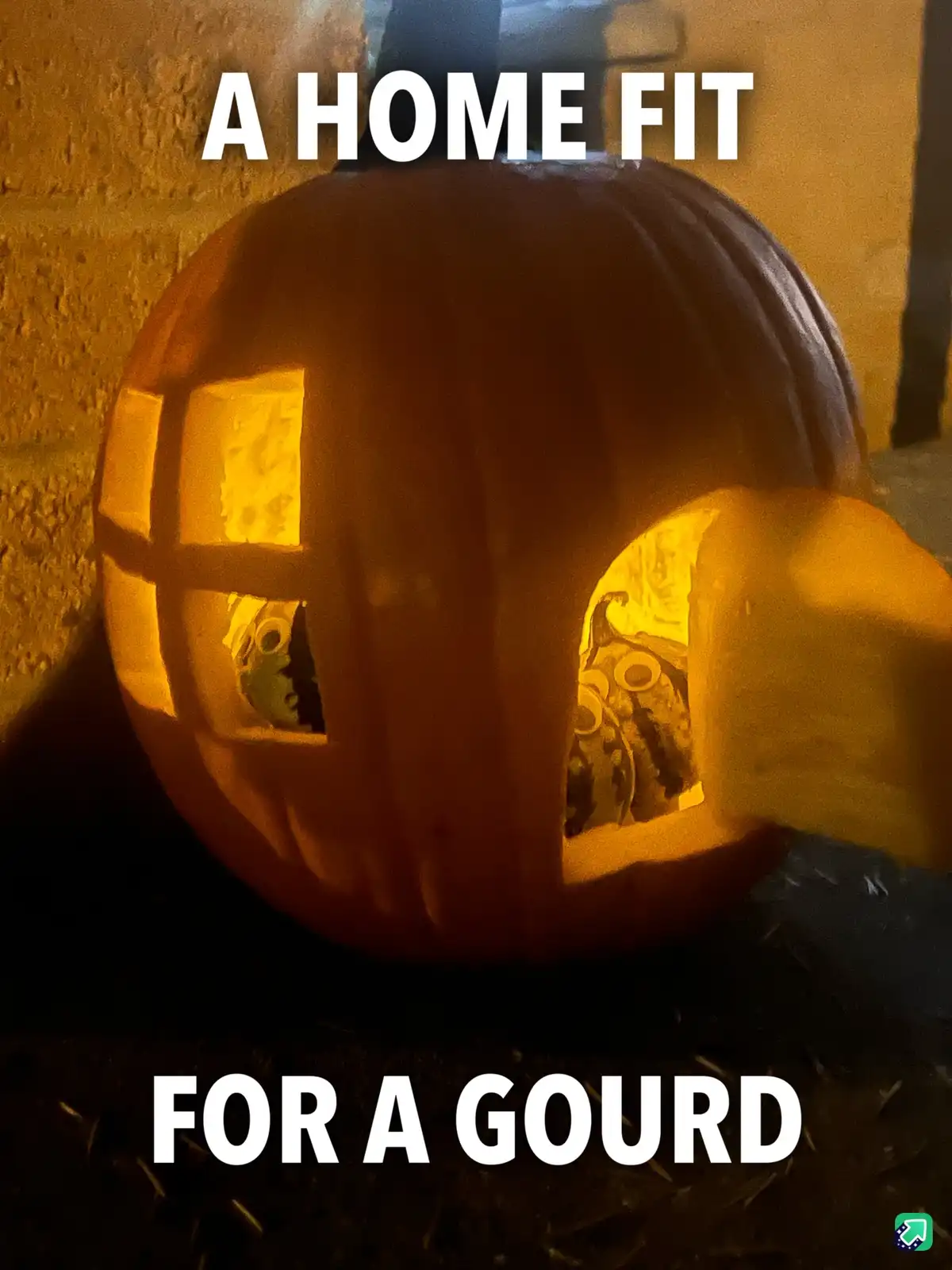 A carved pumpkin with a window and door cut into its side, illuminated from within, sits against a brick wall. The text 'A…