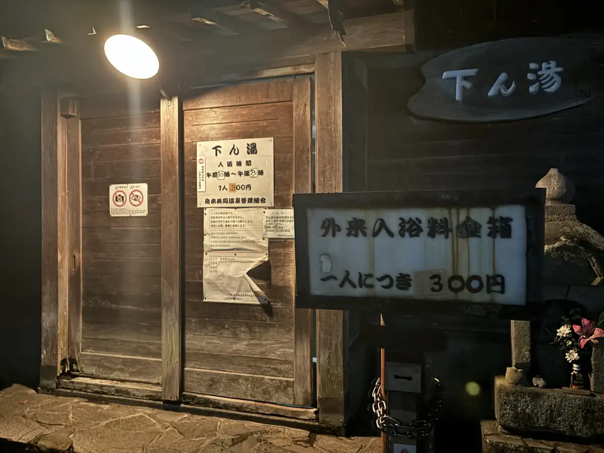 A dimly lit wooden entrance to a traditional Japanese onsen (hot spring) with a sign indicating 300 yen per person for…