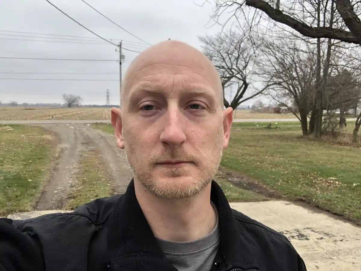 A bald man with a light beard takes a selfie outdoors on an overcast day, standing on a concrete path beside a dirt road…