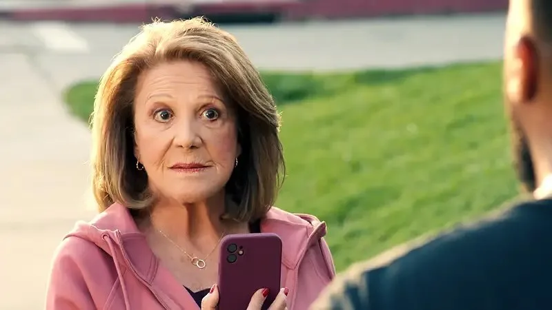 a woman with wide eyes and a slightly downturned mouth holds a purple smartphone while facing a man whose back is to the…