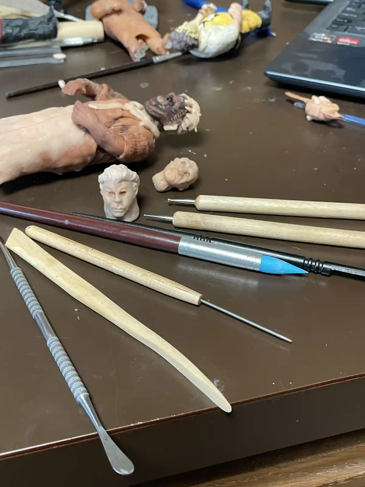 A cluttered artist's desk displays various sculpting tools like knives, brushes, and picks alongside unfinished clay…