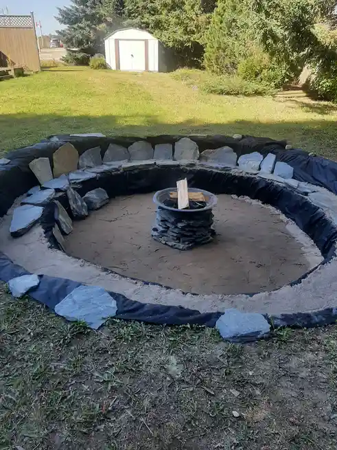 An outdoor fire pit construction site shows a circular depression lined with black plastic sheeting and partially surrounded…