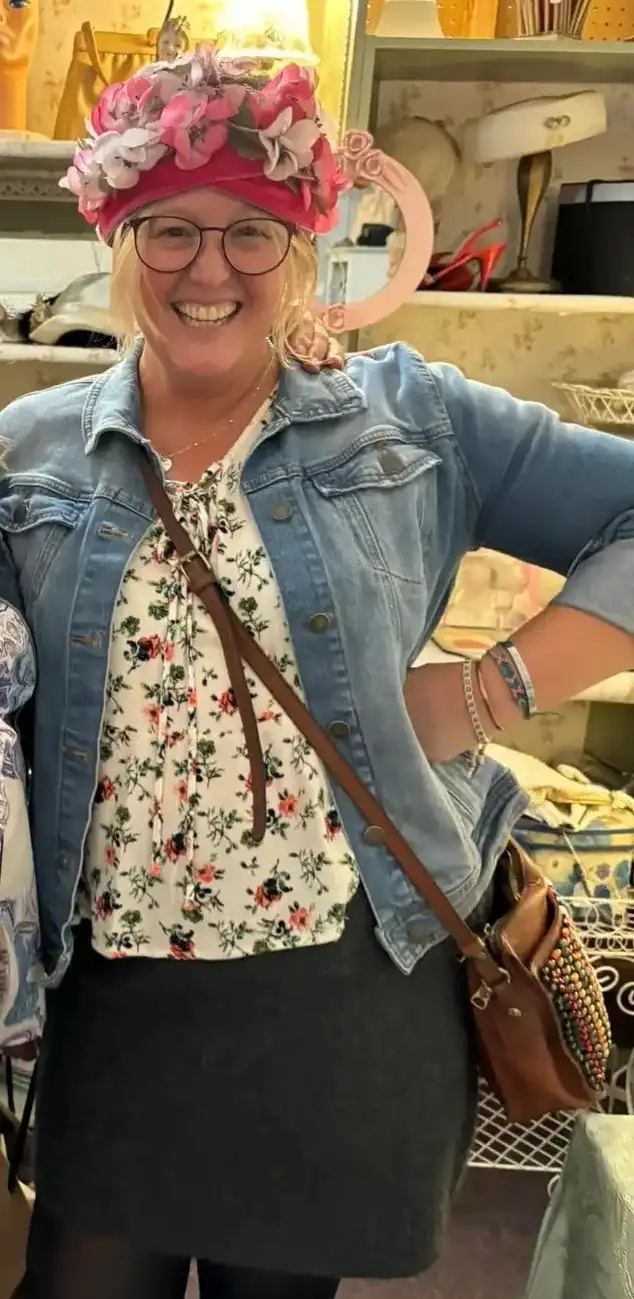 A smiling woman with glasses wears a large, elaborate pink floral hat that covers most of her head, paired with a denim…