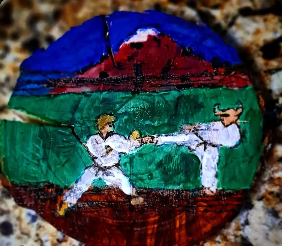 A hand-painted rock depicts two karate fighters in white gi engaged in a martial arts match on a green field, with a…