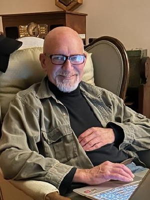 An older, bald man with glasses and a white beard sits comfortably in a chair, smiling while using a laptop. The laptop's…
