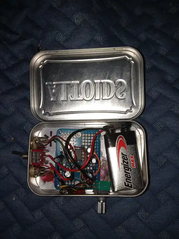 An open silver tin with 'scioTVA' embossed on the lid, revealing a homemade electronic device inside with a blue circuit…