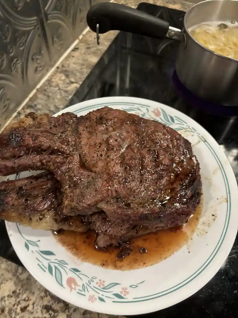 A large, seared tomahawk steak rests on a white plate with a floral pattern, glistening with juices. In the background, a…