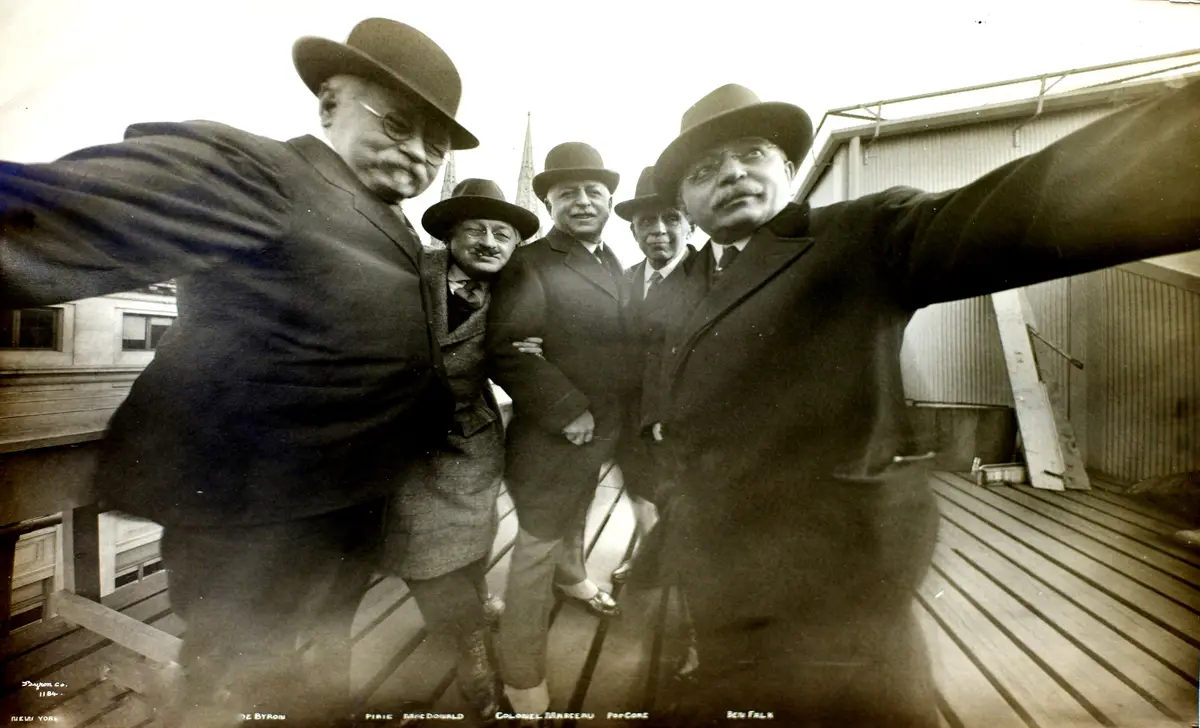 A vintage black-and-white photo shows five men in suits and hats posing on a wooden rooftop, arms outstretched as if taking…