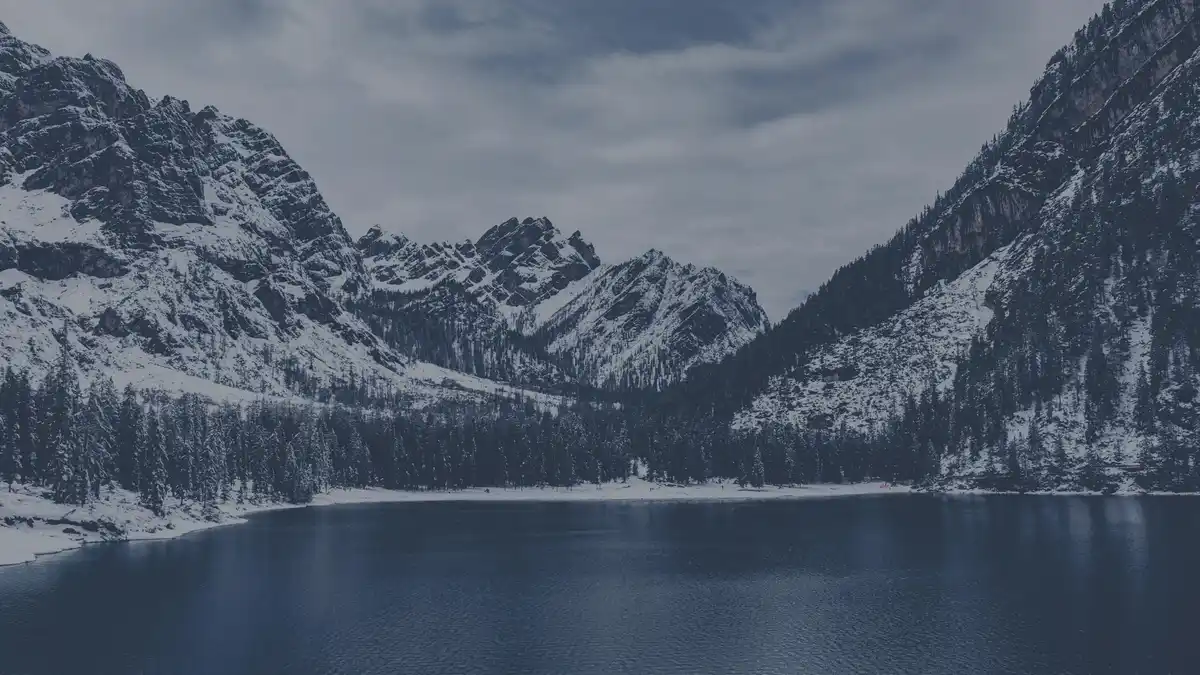 A serene, snow-covered mountain lake surrounded by evergreen forests and rocky peaks under a cloudy sky. The image has a…