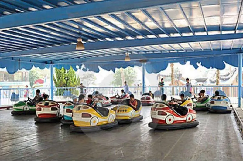 A photo of children riding colorful bumper cars in an amusement park under a blue roof. The cars are small, round, and…