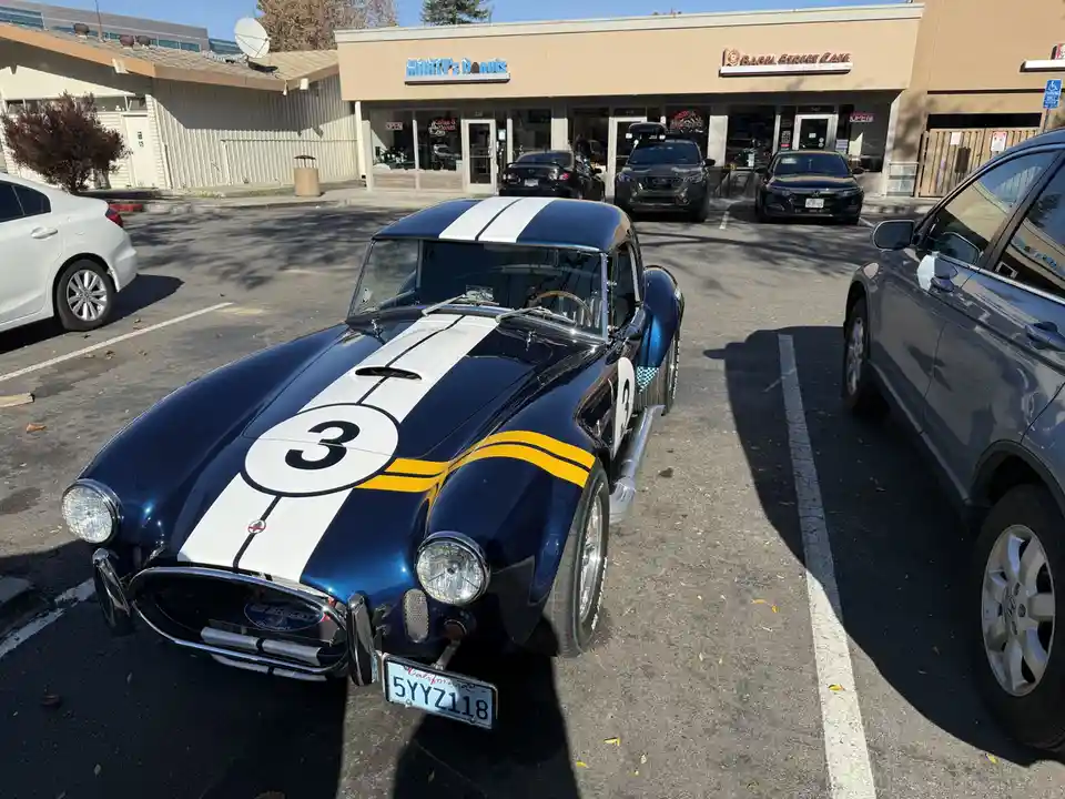 A blue classic Shelby Cobra race car with white and yellow stripes and the number 3 parked in a commercial lot in front of…