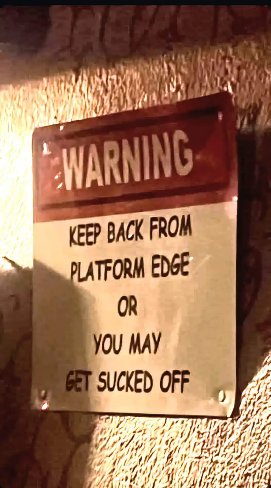 A weathered warning sign mounted on a textured wall reads 'WARNING KEEP BACK FROM PLATFORM EDGE OR YOU MAY GET SUCKED OFF'…