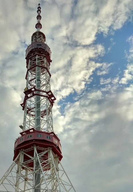 A low-angle photograph shows a tall, red-and-white lattice tower against a partly cloudy blue sky. The tower has multiple…