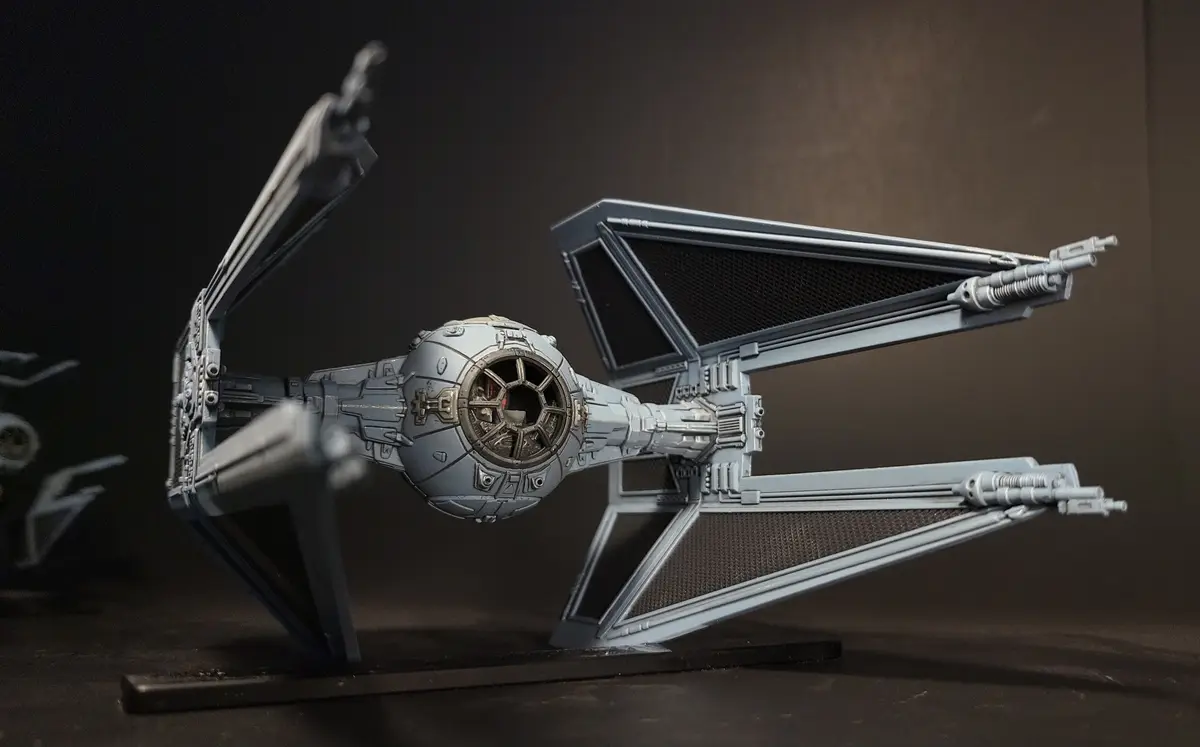 A detailed grey TIE fighter model from Star Wars is displayed on a stand against a dark background. The text overlay reads…