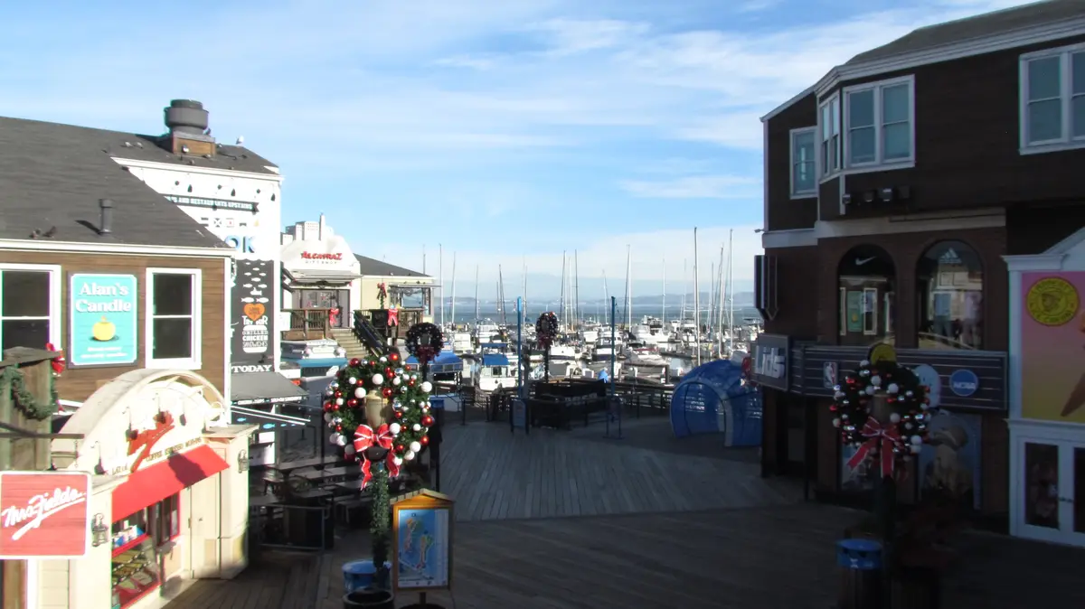 A wide shot of a bustling waterfront boardwalk decorated with large, ornate Christmas wreaths and festive lights. Shops line…
