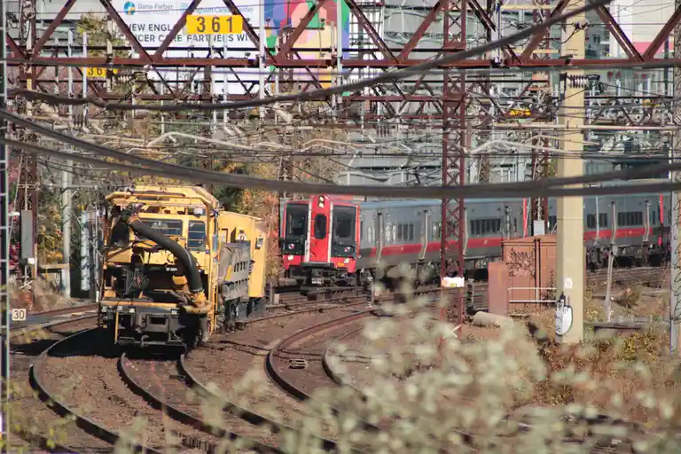 A yellow rail maintenance train is shown on a curved track, facing a red and silver passenger train on an adjacent track…