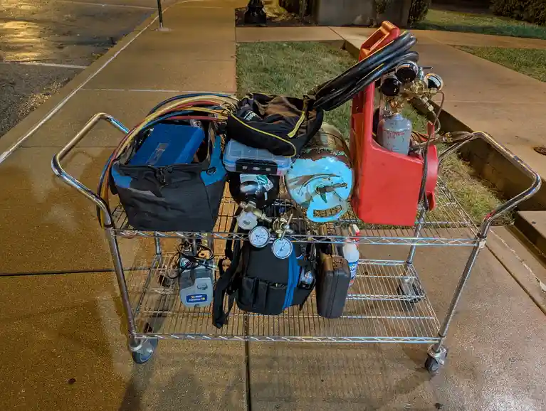 A metal utility cart loaded with HVAC refrigerant equipment, including gas cylinders, gauges, hoses, tool bags, and a vacuum…