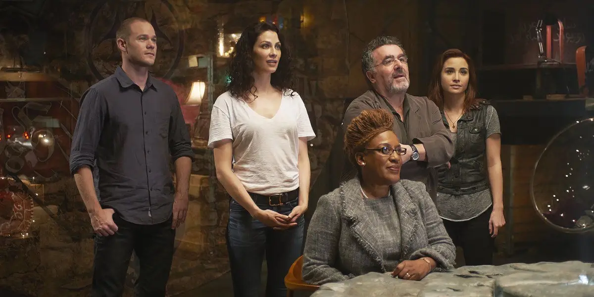 Five people from the TV show 'Warehouse 13' stand in a dimly lit, cluttered room with mystical artifacts. The woman in the…