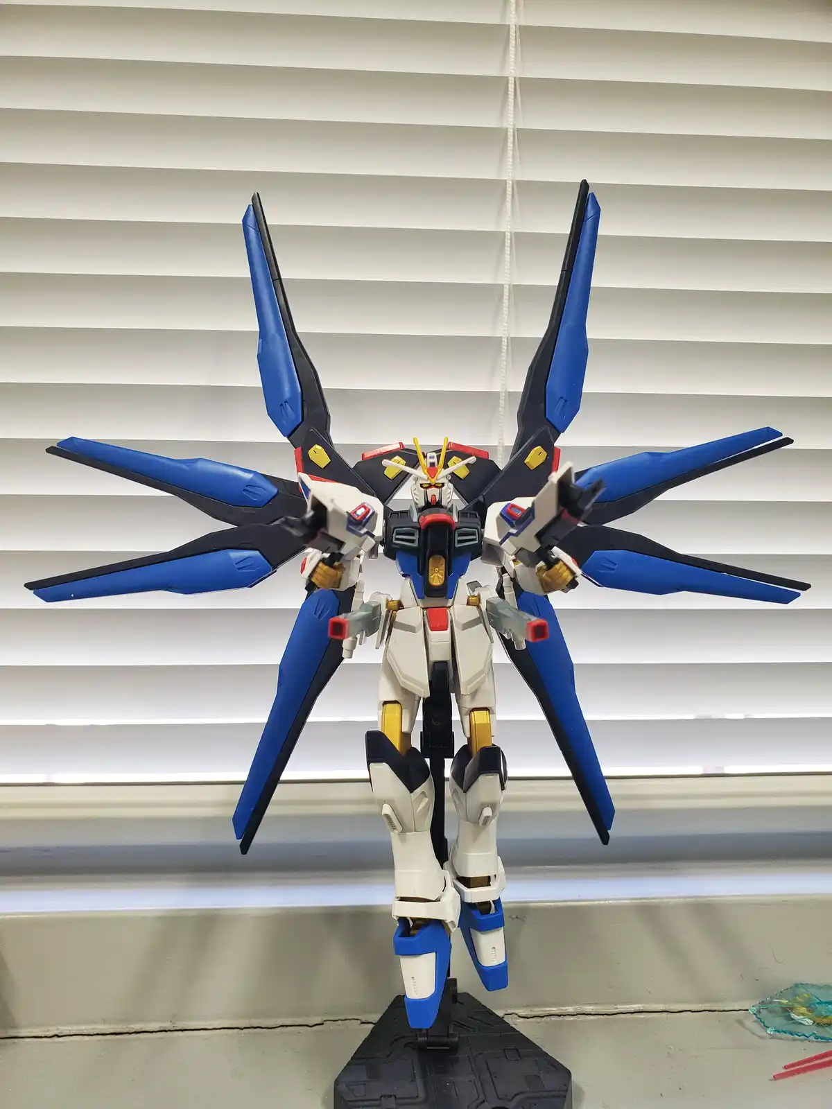 A blue, white, and black Gundam model with large wing-like extensions stands on a display stand against a background of…
