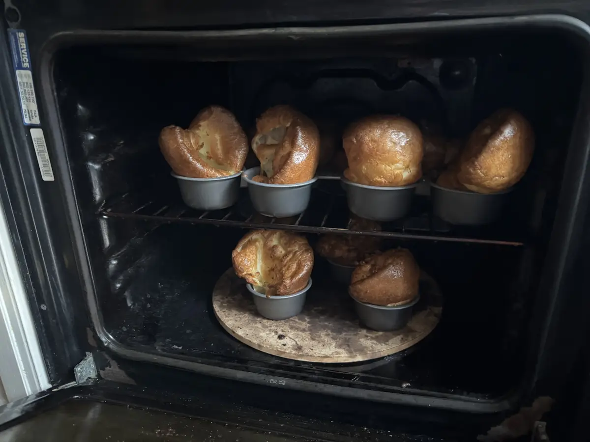 An open oven reveals several golden-brown Yorkshire puddings rising in individual metal cups on two racks. Two of the…