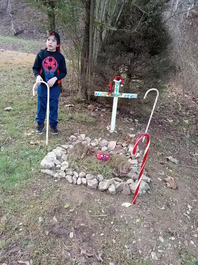 A young boy in a Spider-Man hoodie stands beside a makeshift grave marked by a white cross with '024' and a Santa hat…