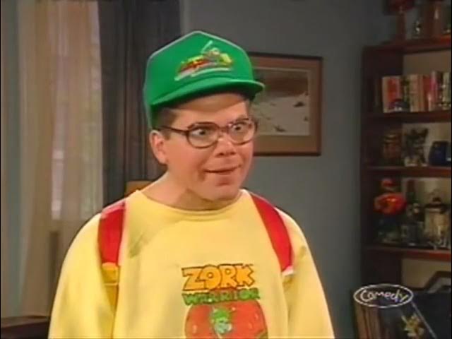 A man wearing a green cap, glasses, and a yellow 'Zork Warrior' t-shirt with red suspenders looks at the camera with a smug…