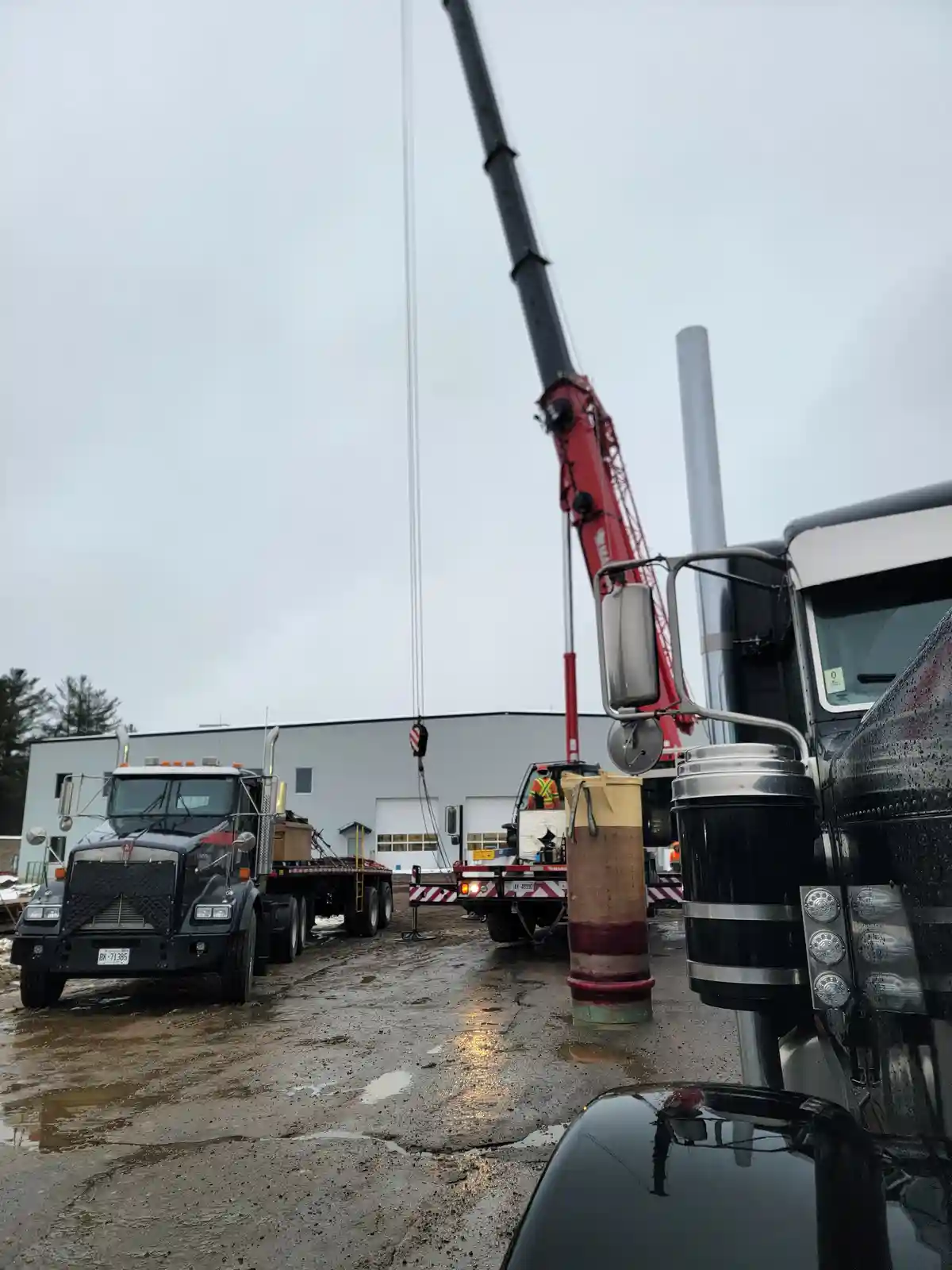 A construction scene with a red crane lifting a large metal pipe, surrounded by heavy trucks on a wet, muddy lot. A worker…
