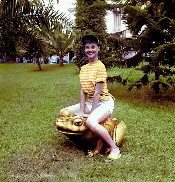 a woman in a yellow striped shirt and white shorts sits atop a golden frog statue in a grassy yard surrounded by palm trees…
