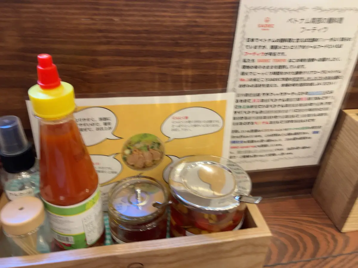 A wooden tray holds condiments including a red sauce bottle, jars of pickled vegetables and dark sauce, and a hand sanitizer…