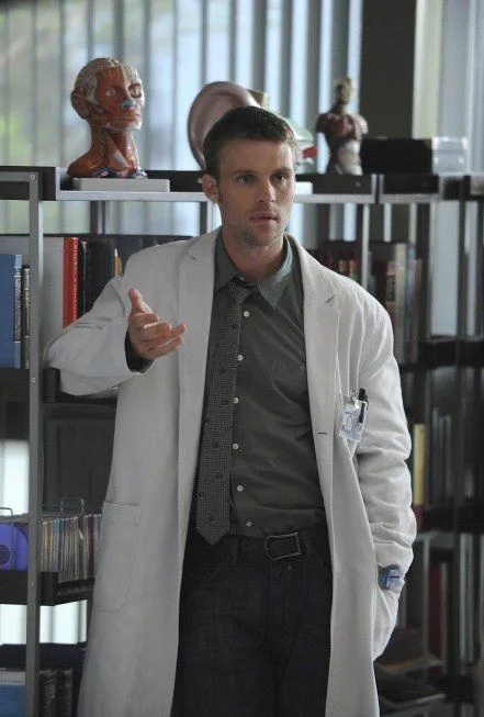 A man in a white lab coat stands in an office with bookshelves and anatomical models behind him, gesturing with his right…