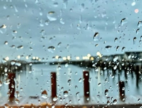 A blurry, out-of-focus photograph taken through a rain-splattered window, showing indistinct city lights and dark vertical…