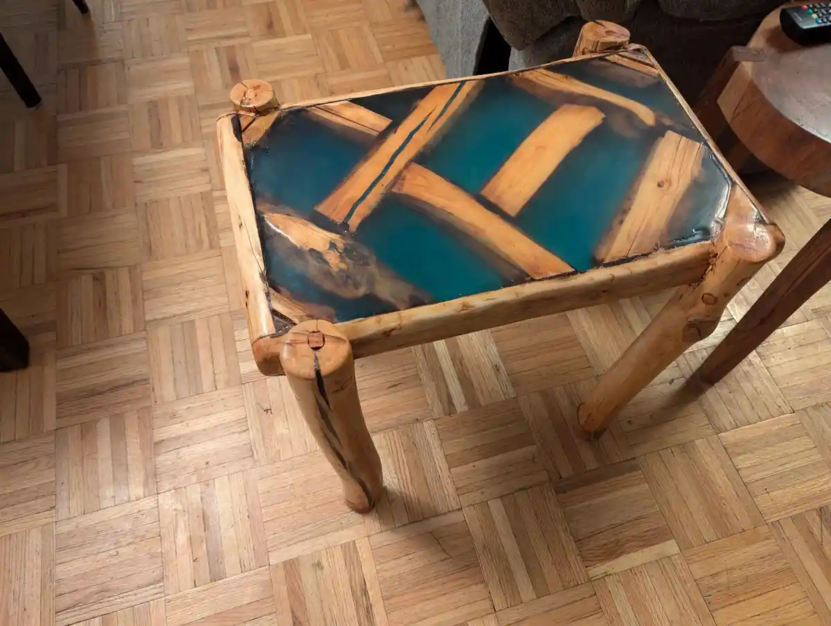 A handmade wooden table with a resin top featuring embedded wood pieces and a blue-green hue, placed on a parquet wood…