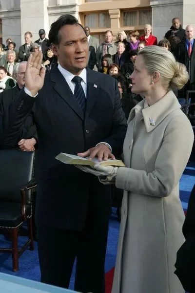 A man in a dark suit and tie raises his right hand while being sworn in, looking upward with a strained expression. A woman…