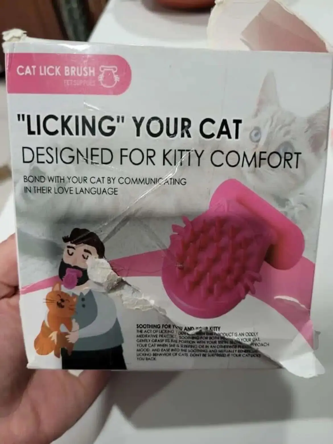 A person holds a product box labeled 'CAT LICK BRUSH' with text claiming it's for 'LICKING YOUR CAT DESIGNED FOR KITTY…