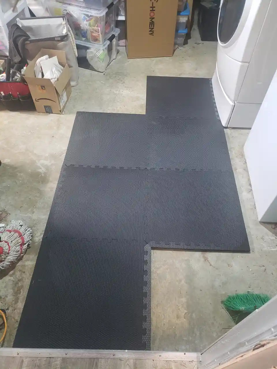 A photo shows four black interlocking foam floor mats arranged in a T-shape on a concrete floor in a cluttered utility room…